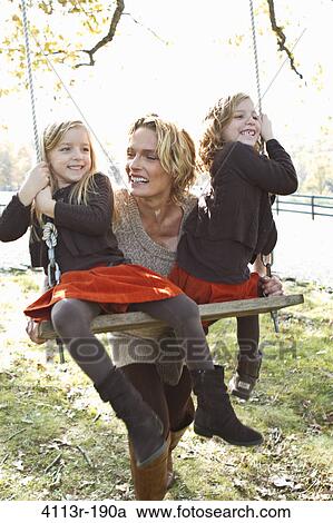 Stock Image - Mother pushing two daughters on swing. Fotosearch
