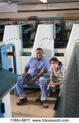 Stock Photo - Portrait of a man and a woman in coveralls sitting in front of a printing machine. Fotosearch