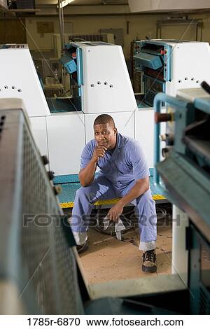 Stock Image - Portrait of a man in coveralls sitting in front of a printing machine. Fotosearch