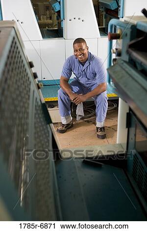 Stock Image - Portrait of a man in coveralls sitting in front of a printing machine. Fotosearch