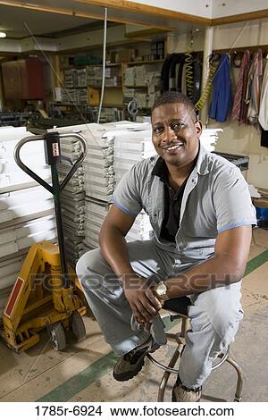 Picture - Portrait of a man sitting on a stool in a warehouse. Fotosearch