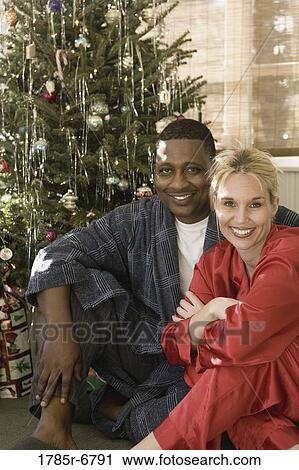 Stock Image - Portrait of a mature interracial couple in pajamas sitting by Christmas tree. Fotosearch