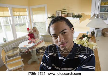 Stock Photography - Portrait of a teenager boy with arms folded, family wrapping gifts in background. Fotosearch