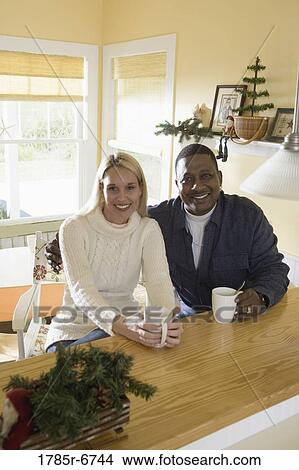 Portrait of an inter-racial couple with coffee mugs sitting at a kitchen counter View Large Photo Image Picture - Portrait of an inter-racial couple with coffee mugs sitting at a kitchen counter. Fotosearch
