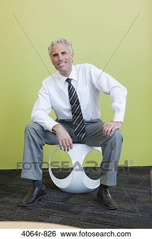 Stock Photograph - Portrait of mature businessman sitting on designer chair against yellow background. Fotosearch