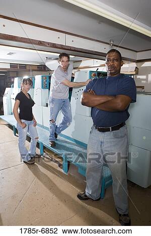 Stock Image - Portrait of two blue-collar men and a woman standing and sitting next two printing machines. Fotosearch