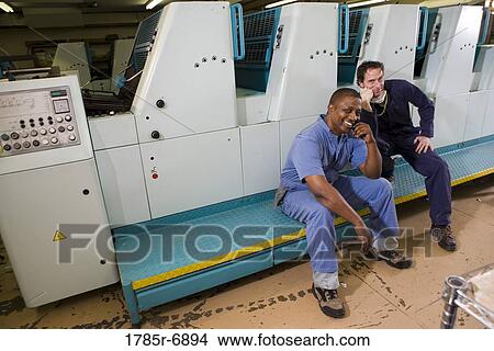 Portrait of two men in coveralls sitting in front of a printing machine View Large Photo Image Picture - Portrait of two men in coveralls sitting in front of a printing machine. Fotosearch