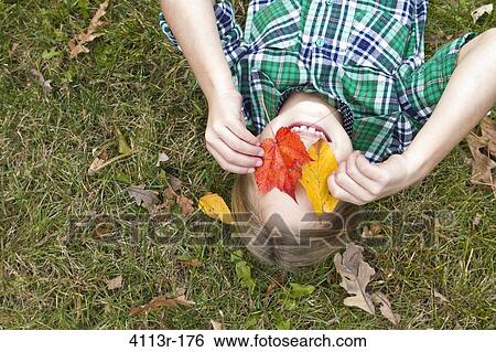 Stock Photograph - Smiling girl holding Autumn leaves in front of face, lying on grass. Fotosearch