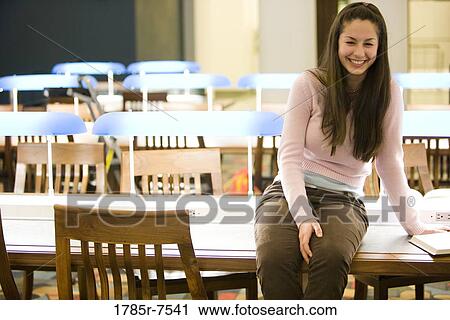Stock Image - Smiling young woman sitting on a desk in the reading room of a library. Fotosearch