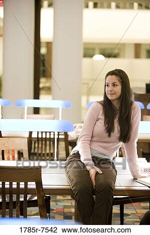 Stock Image - Smiling young woman sitting on a desk in the reading room of a library. Fotosearch