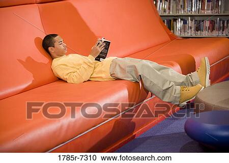 Stock Image - Teenage boy lying on a couch and reading a book in a library. Fotosearch