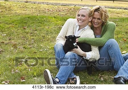 Stock Image - Two women with black lamb sitting on field. Fotosearch