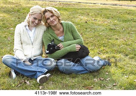 Stock Image - Two women with black lamb sitting on field. Fotosearch