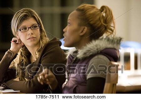 Two young women sitting and conversing in the reading room of a library View Large Photo Image Stock Image - Two young women sitting and conversing in the reading room of a library. Fotosearch