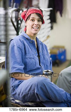 Picture - View of a woman in coveralls sitting down during a coffee break while working in a warehouse. Fotosearch