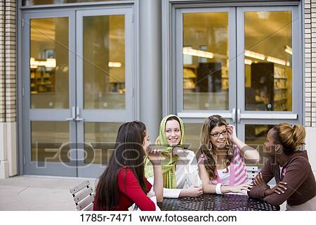 Stock Image - View of four young women conversing while sitting at table outside a library. Fotosearch
