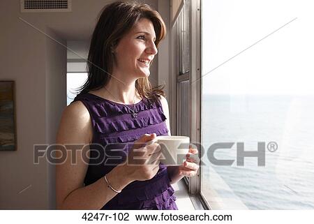 Stock Image - Woman looking at view of water, holding coffee cup and smiling. Fotosearch