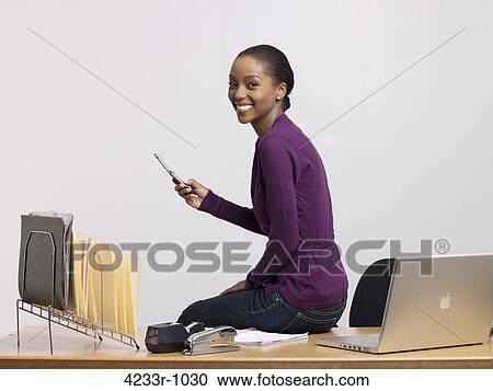 Stock Image - Young businesswoman sitting on desk using phone over colored background. Fotosearch