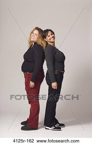 Stock Image - Young female friends standing back to back and smiling. Fotosearch