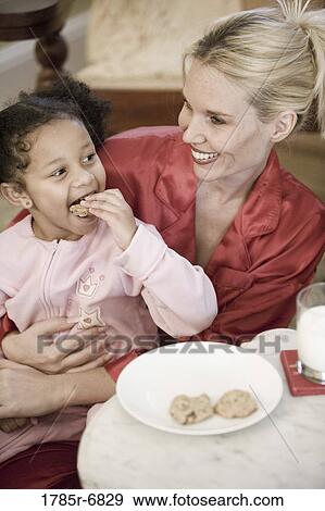 Young girl dressed in pajamas sitting on her mother's lap eating a cookie View Large Photo Image Stock Photo - Young girl dressed in pajamas sitting on her mother's lap eating a cookie. Fotosearch