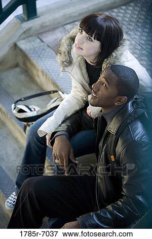 Stock Photo - Young inter-racial couple sitting at the top of a flight of stairs at a public train station. Fotosearch