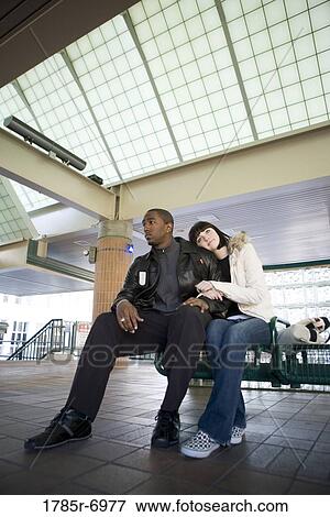 Young inter-racial couple sitting on a bench at a public train station View Large Photo Image Stock Photo - Young inter-racial couple sitting on a bench at a public train station. Fotosearch