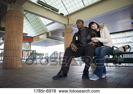 Young inter-racial couple sitting on a bench at a public train station View Large Photo Image Stock Photo - Young inter-racial couple sitting on a bench at a public train station. Fotosearch