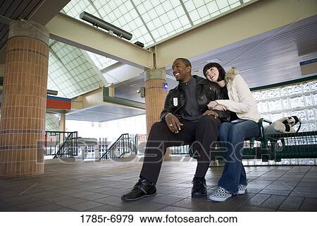 Young inter-racial couple sitting on a bench at a public train station View Large Photo Image Stock Photo - Young inter-racial couple sitting on a bench at a public train station. Fotosearch