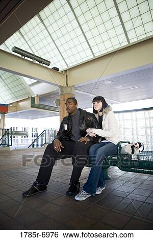 Young inter-racial couple sitting on a bench at a public train station View Large Photo Image Stock Photograph - Young inter-racial couple sitting on a bench at a public train station. Fotosearch