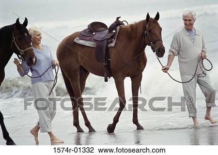 Stock Image - Senior couple walking with horses on the beach. Fotosearch