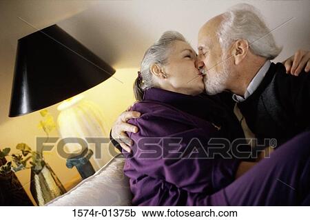 Stock Image - Side profile of a senior couple sitting on a couch kissing each other. Fotosearch