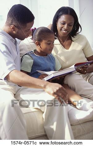Stock Image - Father and mother sitting on a sofa with their daughter reading a book. Fotosearch