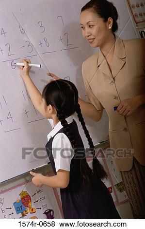 Stock Photo - Girl writing on a board in class with a female teacher standing beside her. Fotosearch