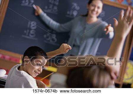 Stock Image - Students with their arms raised and a female teacher standing at the chalkboard. Fotosearch