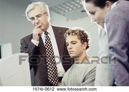 Young man sitting in front of a computer monitor with a professor standing behind him View Large Photo Image Stock Image - Young man sitting in front of a computer monitor with a professor standing behind him. Fotosearch