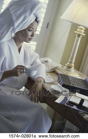 Stock Image - Young woman wearing a bathrobe holding a cup of coffee and looking at a laptop in a hotel room. Fotosearch