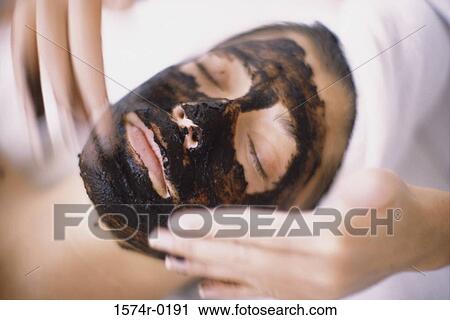 Stock Image - Close-up of a person's hands applying a facial mask on a young woman's face. Fotosearch