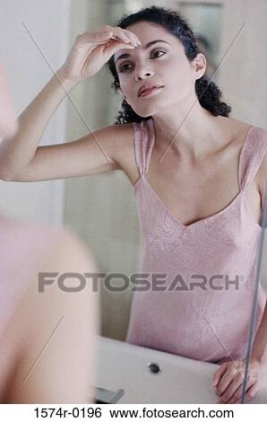 Stock Photograph - Young woman looking in a mirror and tweezing her eyebrows. Fotosearch