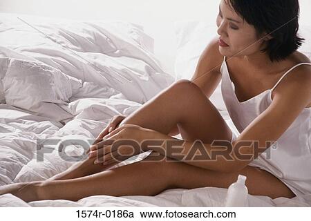 Stock Image - Young woman sitting on a bed and rubbing her leg. Fotosearch