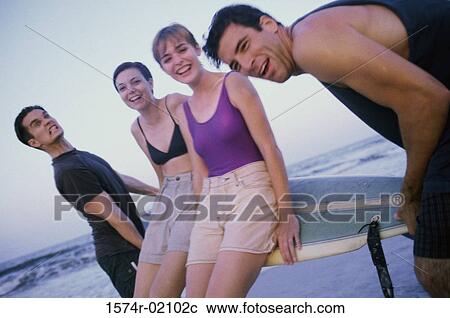 Stock Image - Portrait of two young men carrying two young women on a surfboard. Fotosearch