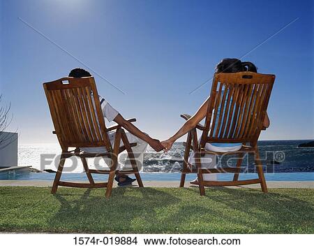 Rear view of a young couple sitting on chairs holding hands View Large Photo Image Picture - Rear view of a young couple sitting on chairs holding hands. Fotosearch
