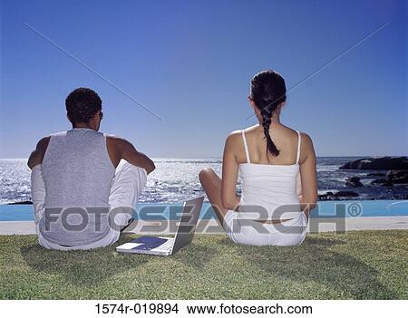 Rear view of a young couple sitting on a lawn with a laptop View Large Photo Image Picture - Rear view of a young couple sitting on a lawn with a laptop. Fotosearch