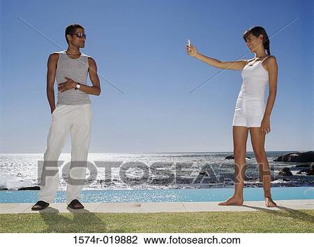 Stock Image - Side profile of a young woman taking a photograph of a young man at the poolside. Fotosearch
