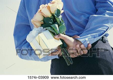 Stock Photo - Rear view of a young man hiding flowers and a gift behind his back. Fotosearch
