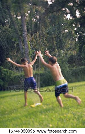Stock Image - Rear view of two boys playing under a water sprinkler. Fotosearch