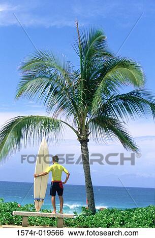 Rear view of a young man holding a surfboard on the beach View Large Photo Image Stock Photograph - Rear view of a young man holding a surfboard on the beach. Fotosearch