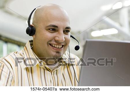 Stock Image - Close-up of a male customer service representative wearing a headset sitting in front of a laptop. Fotosearch