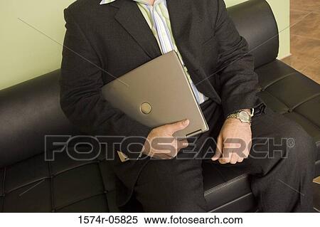 Mid section view of a businessman sitting on a couch holding a laptop View Large Photo Image Stock Photography - Mid section view of a businessman sitting on a couch holding a laptop. Fotosearch
