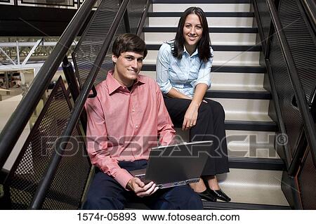 Stock Image - Portrait of a businessman and a businesswoman sitting on stairs in front of a laptop. Fotosearch