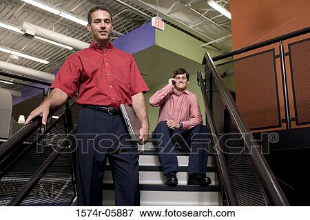 Portrait of a businessman standing on stairs with a businessman sitting behind him talking on a mobile phone View Large Photo Image Stock Photo - Portrait of a businessman standing on stairs with a businessman sitting behind him talking on a mobile phone. Fotosearch